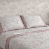 Juego de sábanas Pierre Cardin Paris Jade con bajera rosa lisa, sábana encimera blanca estampada con flores y almohadas a juego con embozo lateral rosa y vivo decorativo, presentado sobre cama con papel pintado neutro.