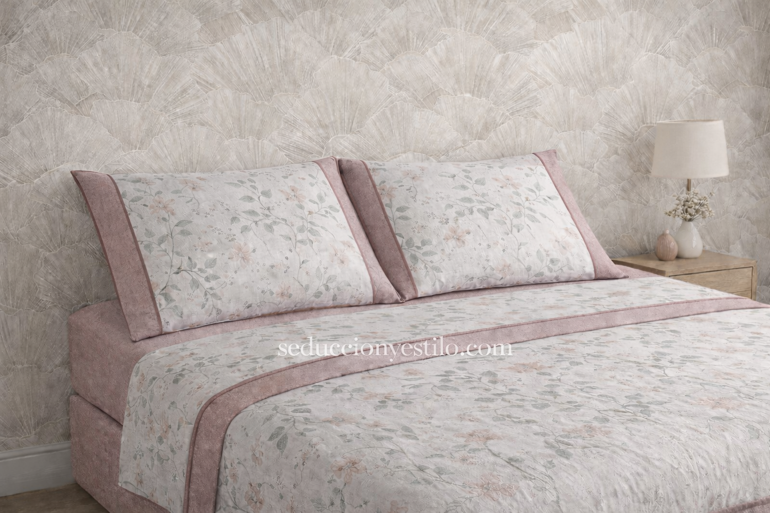 Juego de sábanas Pierre Cardin Paris Jade con bajera rosa lisa, sábana encimera blanca estampada con flores y almohadas a juego con embozo lateral rosa y vivo decorativo, presentado sobre cama con papel pintado neutro.